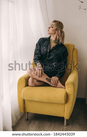 Beautiful young blonde curly woman with makeup and a green wreath on her head in a black shirt sitting, resting on a yellow chair in a white interior interior. Cosiness at home, dreamily girl 