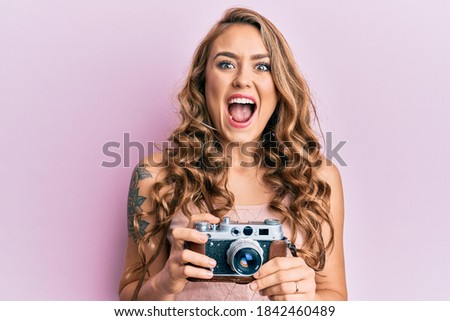 Young blonde girl holding vintage camera celebrating crazy and amazed for success with open eyes screaming excited. 