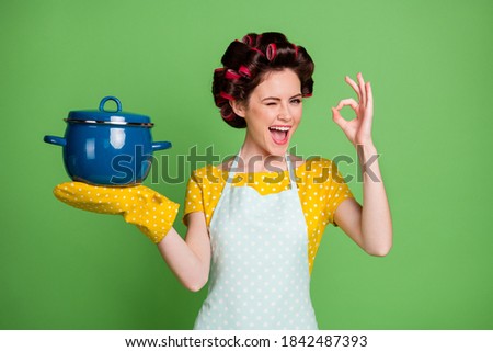Photo of positive cheerful girl wink blink hold saucepan recommend choose decide show okay sign wear yellow dotted t-shirt hair rollers pot holder isolated over green color background