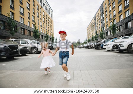 Boy and a girl are running along the city street. A blonde with ponytails in a pink dress and a guy in a baseball cap among cars and buildings.