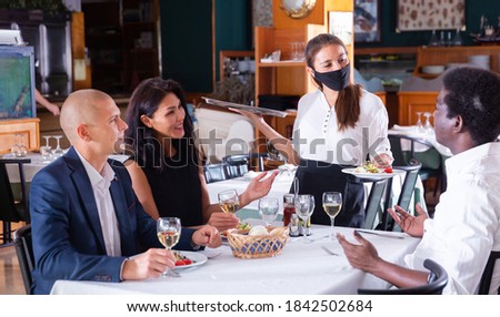 Hospitable waitress wearing face protective mask bringing ordered meals to guests. New normal restaurant concept in coronavirus pandemic .. Royalty-Free Stock Photo #1842502684