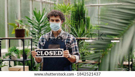 Portrait of young happy Caucasian male owner of garden center holding Closed sign and looking at camera. Handsome flower shop worker in apron and mask standing alone indoors. Job concept