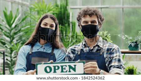 Close up of young Caucasian male and female joyful owners of garden center holding Open sign and smiling to camera. Cheerful flower shop workers in masks posing to camera indoors. Business concept