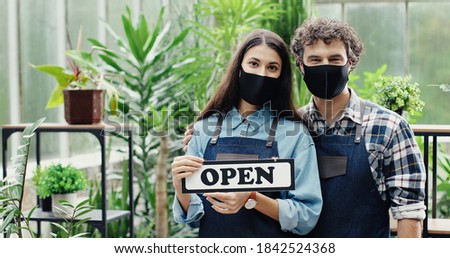 Close up portrait of happy young Caucasian male and female florists holding Open sign with smile on faces. Joyful flower shop employees in masks posing to camera indoors. Floral business concept