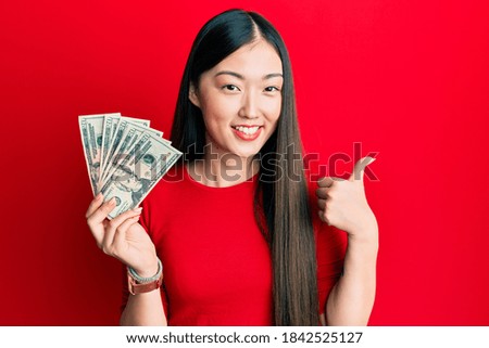 Young chinese woman holding 20 dollars banknote smiling happy and positive, thumb up doing excellent and approval sign 
