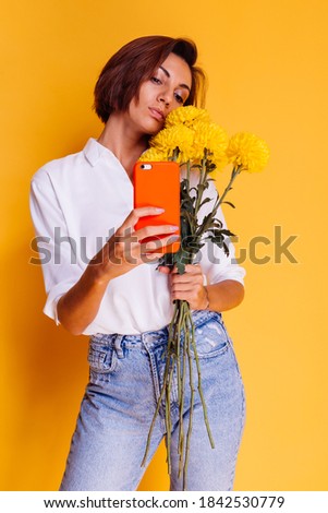 Happy caucasian blogger woman in casual clothes holding yellow asters from flower shop, studio shot, taking photo selfie in mirror on mobile phone for social media content. 