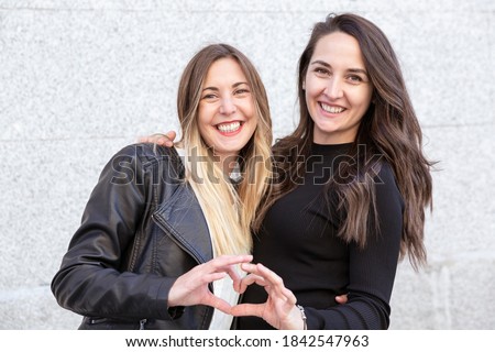 Two smiling girls hugging and forming a heart with their hands. They are out in the open.