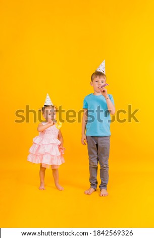 a small boy and a girl stand tall in holiday hats and blow whistles on a yellow background with a copy of space