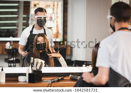 mirror reflection of barber in protective equipment near client in medical mask, blurred foreground Royalty-Free Stock Photo #1842581731