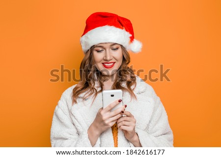 Happy caucasian woman in christmas hat and white faux fur coat holding mobile phone in her hands, posing in studio and texting isolated on orange background