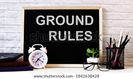 GROUND RULES is written on the chalkboard next to the white alarm clock, glasses, potted plant, and pencils in a stand.