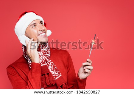 Smiling man in santa hat holding sparker while talking on smartphone isolated on red