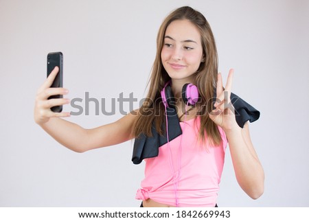 Beautiful smiling fit sportswoman taking a selfie and showing peace gesture while standing isolated over white background