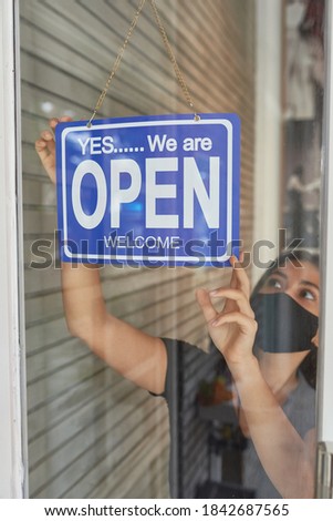 Beautiful European girl in a mask hangs a sign on a glass door of a clothing store (open) after a cowid-19 epidemic