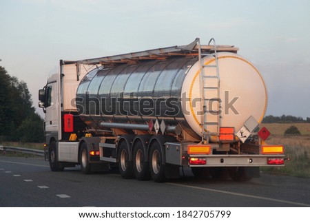 Three-axle semi truck with shiny chrome barrel tank with clear place on ADR sign moving on empty suburban asphalt highway road on a summer day on blue sky, dangerous cargo logistics, back side view