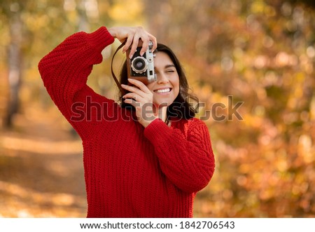 Portrait of cheerful female photographer taking photos of beautiful autumn with retro camera at autumn park. Lovely millennial lady photographing bright nature on warm fall day