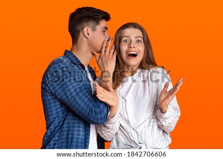 Secrets and gossip of friends. Young man whisper in female ear something amazing and shocked woman with open mouth gestures and look at camera isolated on orange background, studio shot, empty space
