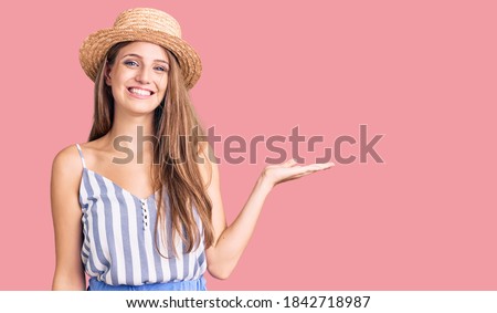 Young beautiful blonde woman wearing summer hat smiling cheerful presenting and pointing with palm of hand looking at the camera. 