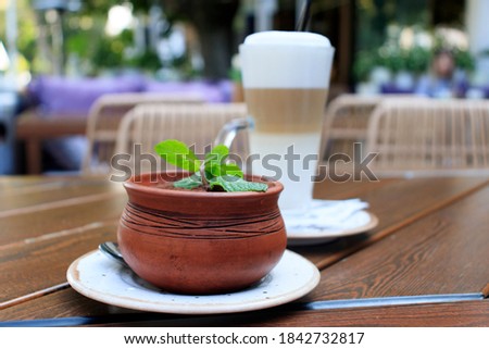 A Cup of hot latte and tiramisu dessert with a sprig of mint in a clay pot on a wooden table in a street coffee shop on a blurry background.