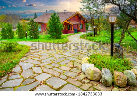 A wooden cottage with a green lawn. Landscape design. Stone-paved paths, lanterns and trees near the cottage. House in a beautiful location. Country vacation in autumn.