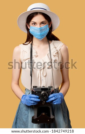 Travel photo. Covid-19 protection. Summer trip. Holiday memories. Woman in medicine mask gloves holding retro camera looking straight isolated on beige.