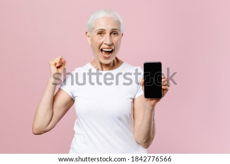 Excited elderly gray-haired female woman in white t-shirt hold mobile phone with blank empty screen mock up copy space doing winner gesture isolated on pastel pink color background studio portrait