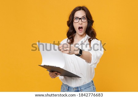 Shocked young brunette business woman in white shirt glasses isolated on yellow background studio. Achievement career wealth business concept. Mock up copy space. Hold clipboard with papers document