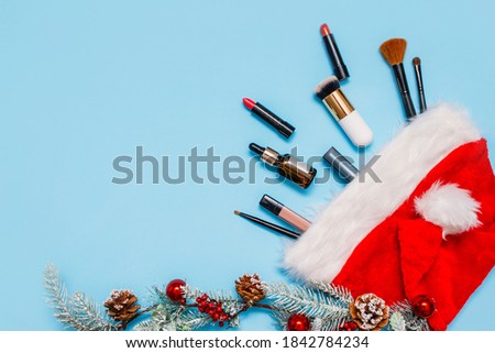 Flatlay with makeup products and Christmas decor on a blue background. make-up brushes, lipsticks, and a jar of oil protrude from Santa's red hat . The view from the top. the concept of women's wishes