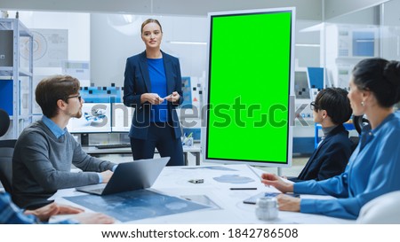 Modern Industrial Factory Meeting: Confident Female Engineer Uses Interactive Green Mock-up Screen Whiteboard Template in Vertical Mode to Make a Report to a Group of Engineers, Managers