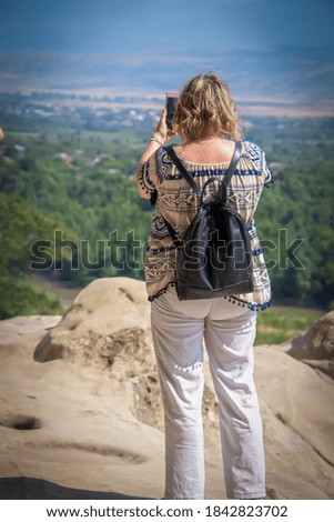 Overweight woman tourist with purse backpack taking a selfie with a pink phone standing on rock outcrop overlooking a wide forested valley with mountains in the background.