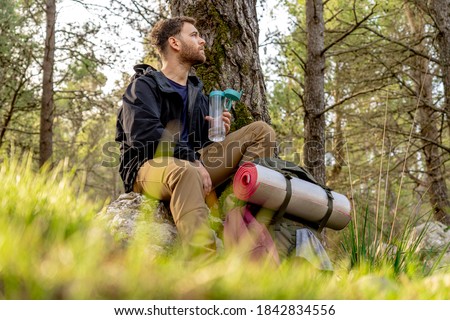 Young hiker sitting on a stone observing the vast mountain forest recovering energy, man wears camping clothes and large backpack