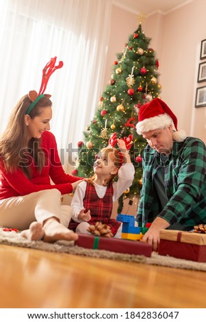 Beautiful young family sitting by nicely decorated Christmas tree, having fun while opening Christmas presents, parents playing with their daughter