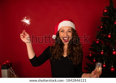 Cheerful attractive woman in santa hat celebrating New Year, Christmas, Hanukkah, Kwanzaa holiday. Smiling lady with sparkler on red festive background. Holiday promotions discounts offers xmas sale