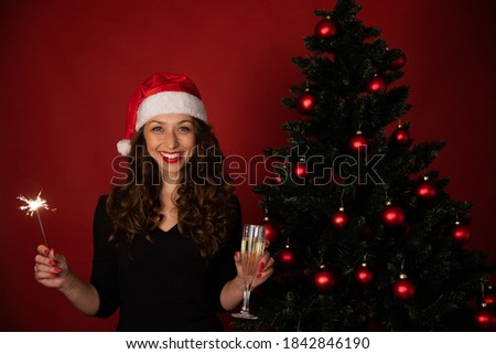 Cheerful charming woman in santa hat celebrating New Year, Christmas, Hanukkah, Kwanzaa holiday. Smiling lady with sparkler and champagne on red background. Xmas sale promotions, congratulation