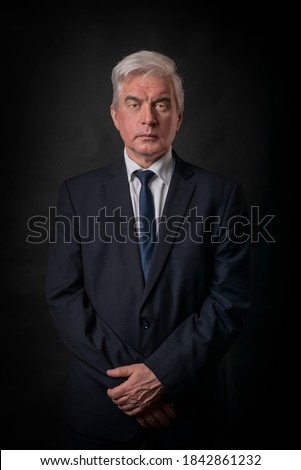 A mature, rich and stylish gray-haired man and gentleman, a businessman, a professor in a dark suit and tie, who looks with an attentive and piercing gaze and with crossed arms.