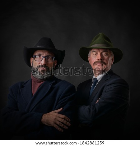 Two mature men, rich gentlemen with beards and mustaches, in dark clothes, coats and suits and hats, against a dark or black background, who look ahead with an attentive and piercing gaze.
