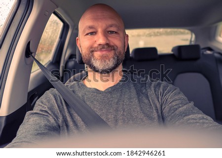 Bald male driver in focus, steering wheel out of focus. Man wears grey shirt, unshaven with grey and black beard in his 40s. Toned. Smiling expression on his face