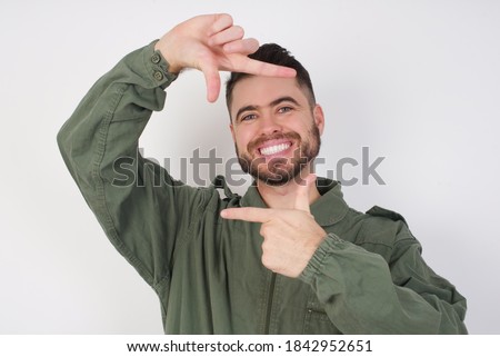 Young caucasian male wearing green overall isolated over white background making finger frame with hands. Creativity and photography concept.