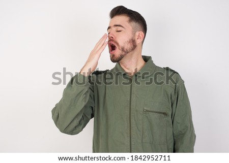 Young caucasian male wearing green overall isolated over white background being tired and yawning after spending all day at work.