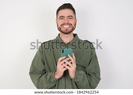 Young caucasian male wearing green overall isolated over white background enjoys distant communication, uses mobile phone, surfs fast unlimited internet, has pleasant smile