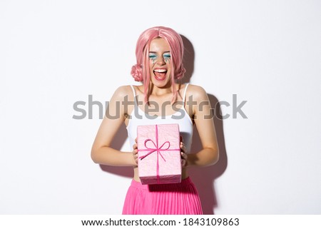 Cheerful birthday girl looking excited, wearing pink wig, shouting of joy, receiving bday gift, standing over white background and celebrating