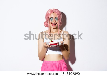 Image of flirty girl in pink wig, showing tongue from temptation, holding delicious piece of cake, standing over white background