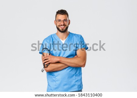 Healthcare workers, medicine, covid-19 and pandemic self-quarantine concept. Cheerful handsome doctor in blue scrubs and glasses, cross hands confident, hold stethoscope and smiling