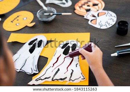 Over shoulder view of unrecognizable child sitting at table and applying paint on ghost picture while making Halloween decoration