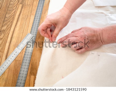 designer's hand fixes paper cutting layouts of dress to calico fabric by pins on wooden table at home