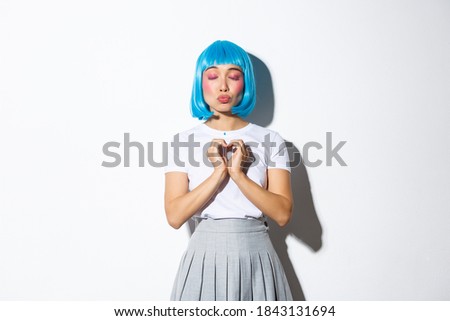 Portrait of lovely asian girl in blue wig and halloween costume, close eyes and pouting while showing heart gesture, daydreaming, standing over white background