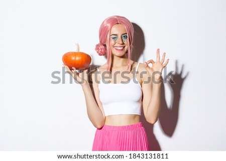 Image of happy girl celebrating halloween, wearing pink wig, holding pumpkin and showing okay sign, standing over white background