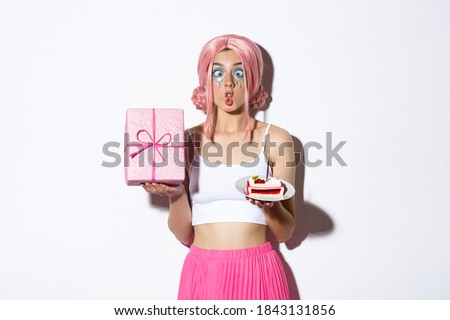 Image of funny birthday girl making silly faces, holding b-day cake and wrapped gift, standing over white background