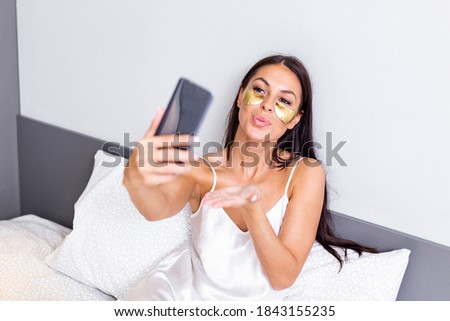 Young happy woman with Gold Cosmetics mask, Eye Patches using mobile phone for video call , gesturing kisses to friends. Beautiful girl making facetime video calling with smartphone lying on the bed.