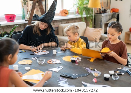 Concentrated young art teacher in witches hat sitting at table and drawing Halloween pictures with kids while preparing paper decorations
